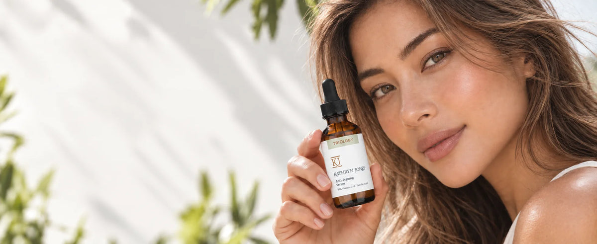 A woman outdoors holding a KJ Serums Triology Vitamin C serum bottle next to her face, against a plain light background.