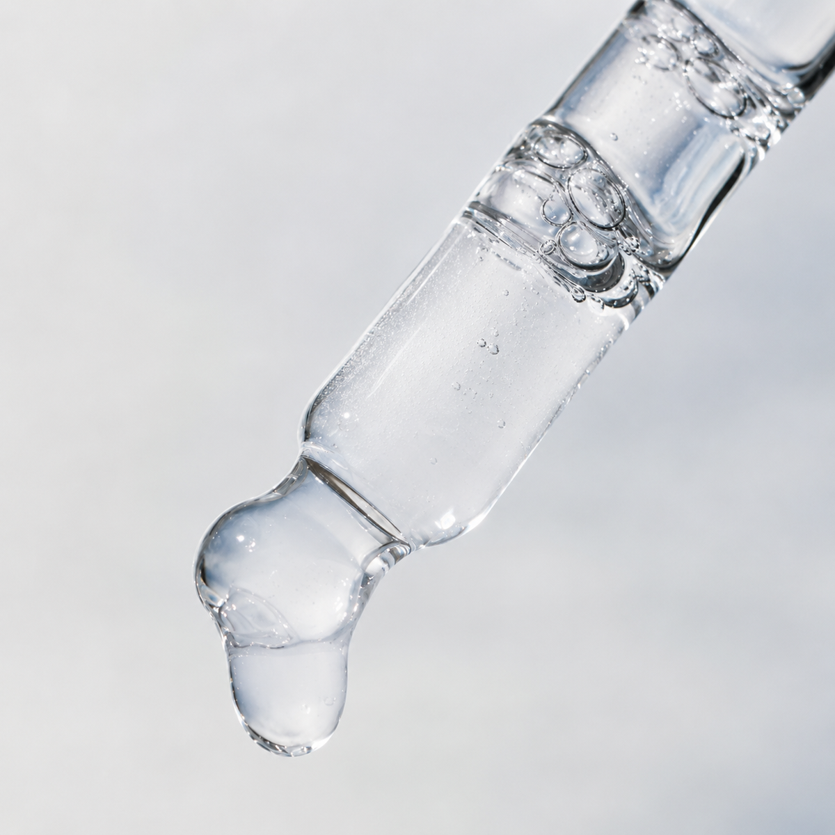 A close-up of a clear glass dropper containing cloudy liquid, with a single glossy drop hanging at the tip against a soft light gray background.