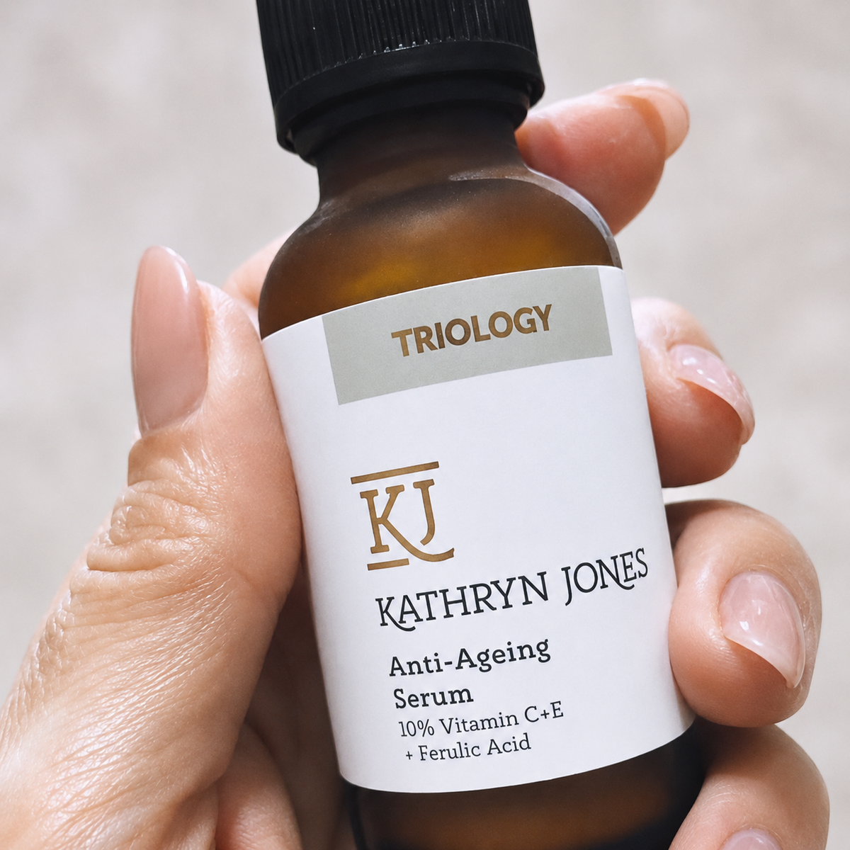 A hand holding a small amber glass bottle of Kathryn Jones Triology Anti-Ageing Serum with a white label and black cap against a soft neutral background.